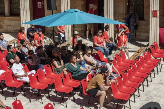 Le public sur des chaises rouges se protège du soleil sous un parasol bleu.