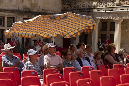 Le public, assis sur des chaises rouges s'abritent du soleil sous un parasol dans la cour du collège