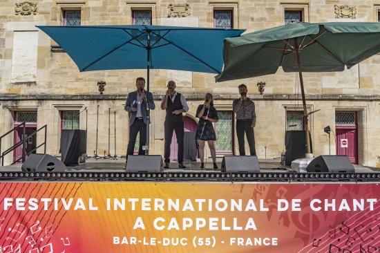 Photo de groupe, sur la scène du collège Gilles de Trèves, des 4 chanteurs de Climax micro à la main et abrités du soleil par deux parasols. en avant plan, la banderole du festival
