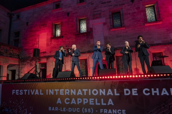 Les 6 membres du groupe Slixs en train de chanter devant un mur éclairé de mauve. EN avant plan, la banderolle du festival