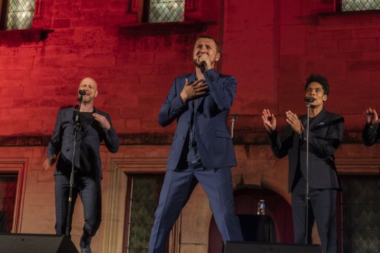 3 membres du groupe Slixs en train de chanter devant un mur éclairé d'orange et de rouge.