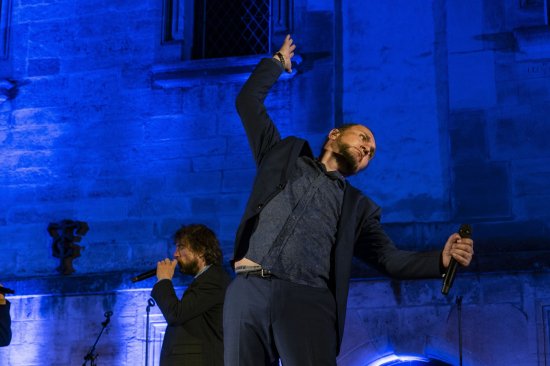 Un chanteur du groupe Slixs se penche en arrière, les bras au ciel. en arrière plan, un chanteur micro à la main. Le mur de fond est éclairé de bleu.