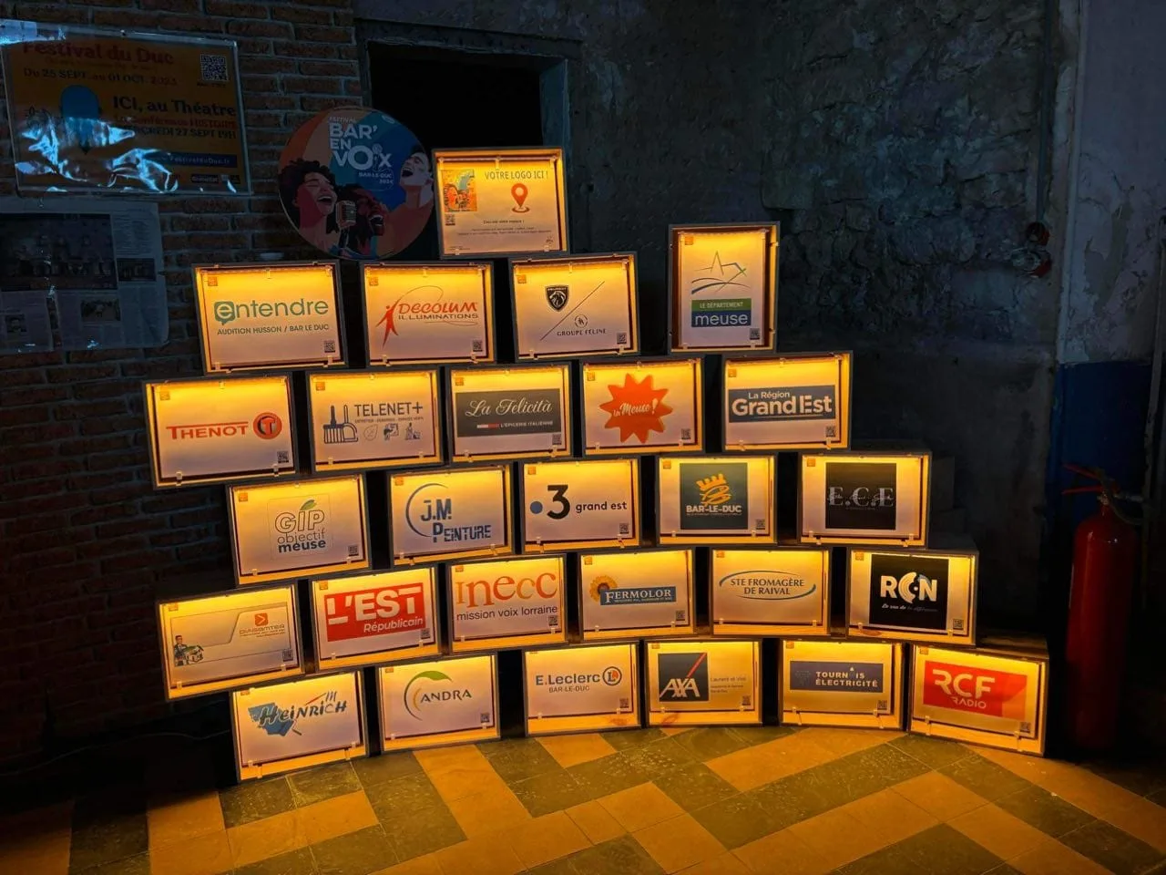 Mur de caisses de vin en bois éclairées, avec le logo des partenaires du festival.