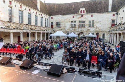 Le public assis sur des chaises rouges, écoute attentivement les artistes sur la scène du collège Gilles de Trèves sous un ciel menaçant.