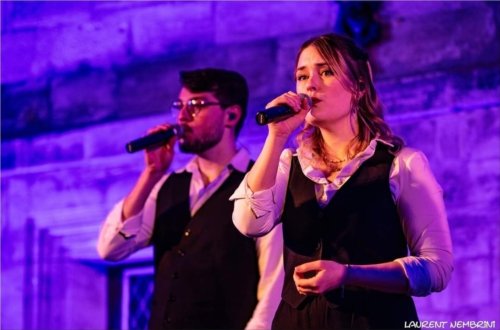 Oriane et Philippe du groupe Just Vox, micro à la main en train de chanter devant une façade de pierre éclairée de bleu et mauve.