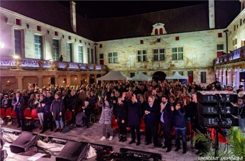 Le public applaudit, debout, les artistes dans une cour du collège Gilles de Trèves illuminé de blanc et mauve dans la nuit tombée.
