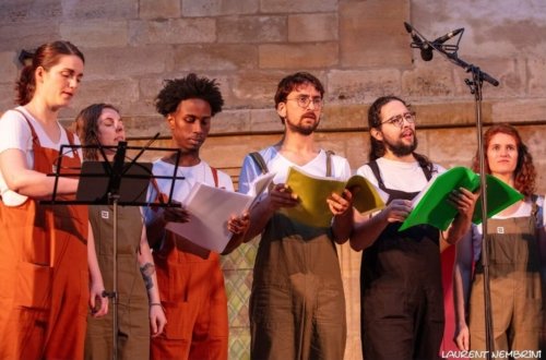 3 chanteurs et chanteuses de l'Atelier Chorale U derrière un micro, en salopettes de toutes les couleurs et partitions à la main en train de chanter.