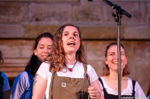 Une chanteuse de l'Atelier Chorale U en gros plan derrière un micro. Deux autres chanteuses en arrière-plan.