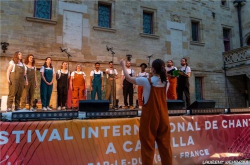 Un membre de l'atelier Chorale U dirige, depuis la cour du collège, les autres membres du groupe qui sont sur scène. La nuit est en train de tomber.