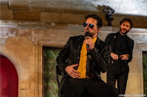 Andrea du groupe Mezzotono, assis sur une chaise, lunettes noires et micro à la main est en train de chanter. Dans le fond, Marco l'observe