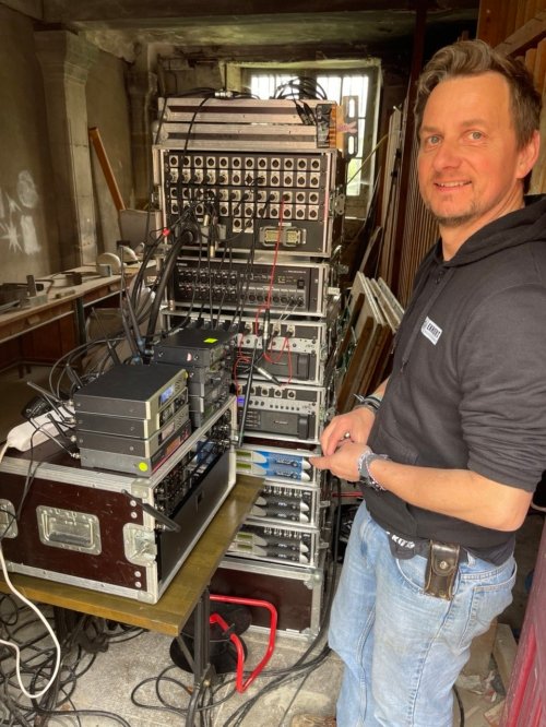 Nico, souriant devant des racks d'amplis, préamplis, splitters et autres équipements de sonorisation.