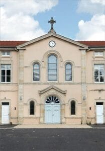 Façade de la chapelle du Lycée Raymond Poincaré avec 3 vitraux et une horloge surmontée d'une croix de pierre.