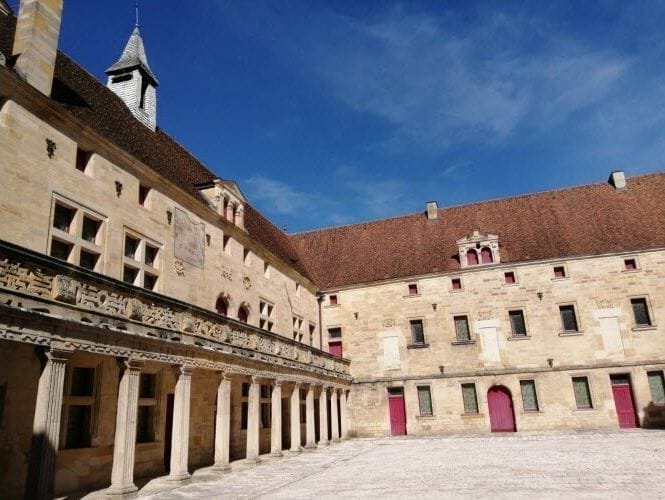 Vue ensoleillée de la cour Gilles de Trèves, patrimoine de Bar-Le-Duc avec les coursives à gauche portées par des colonnes de pierre et le bâtiment de fond de cour, sous un ciel bleu.