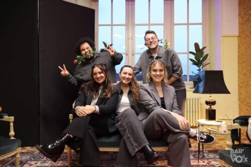 Photo de groupe de JustVox dans les loges. Les filles assises à l'avant, les garçons, debout à l'arrière avec une rose en bouche
