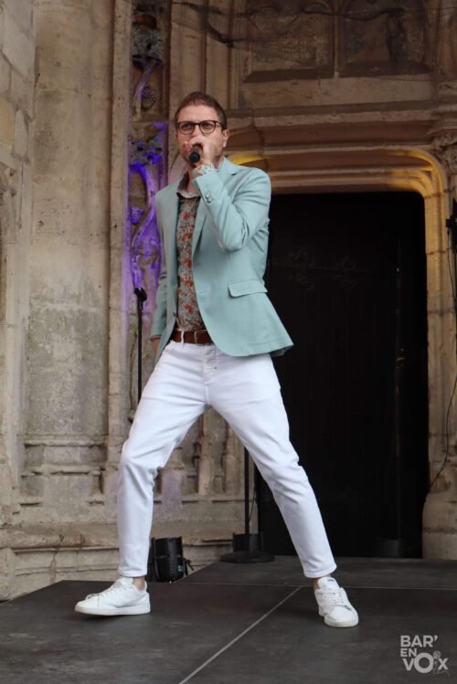 Paolo, en veste verte et pantalon blanc chante micro à la main
