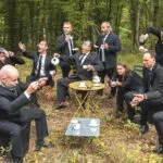 Les 12 chanteurs de VoxpoP, assis autour d'une table dans la forêt, s'amusent.
