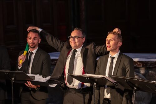 trois chanteurs, celui du milieu tient ses collègues par les cheveux en souriant